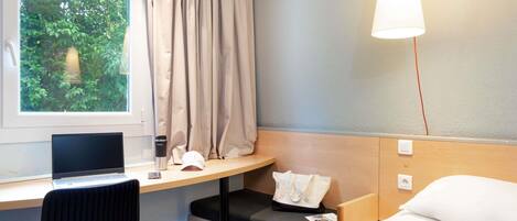 A hotel room with a bed, a desk with a laptop, a chair, a window with curtains, and a hanging light fixture.