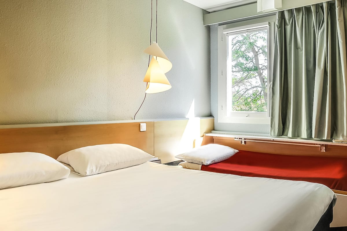 Habitación de hotel con cama, mesita de noche, lámpara, ventana con cortinas y vistas a árboles.