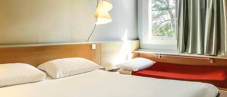 A hotel room with a bed, a bedside table, a lamp, a window with curtains, and a view of trees outside.