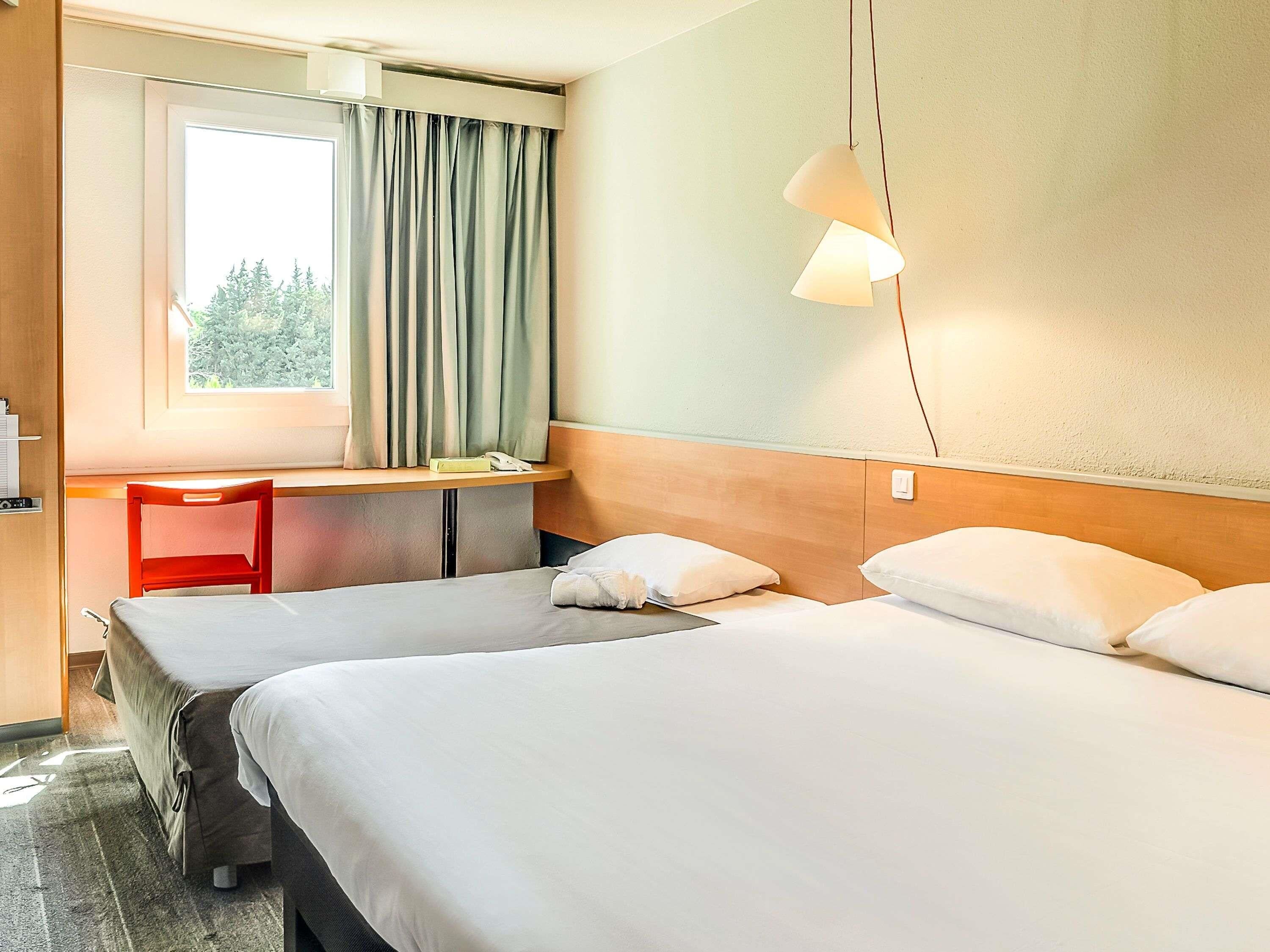 Une chambre d’hôtel avec deux lits, un bureau, une chaise et une fenêtre avec des rideaux.