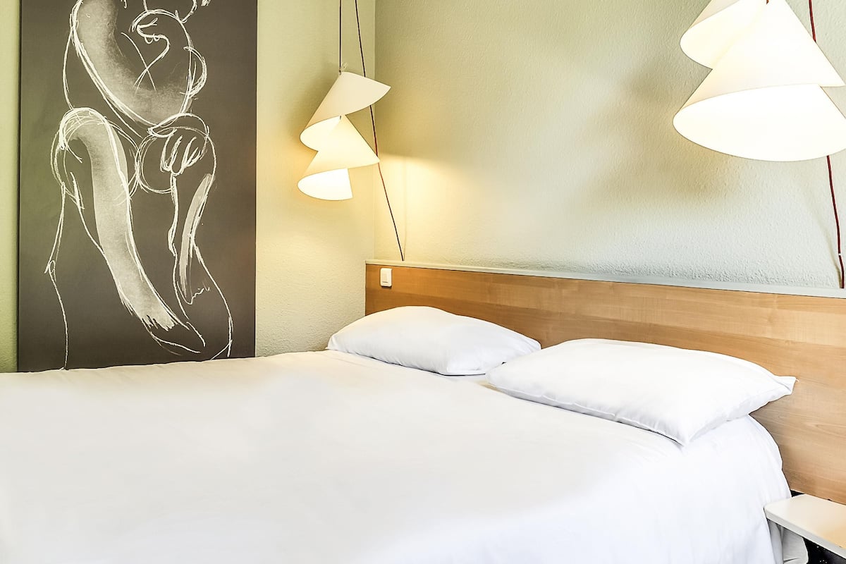 Una habitación de hotel con una cama, almohadas blancas, un cabecero de madera, una obra de arte en la pared y dos lámparas colgantes.