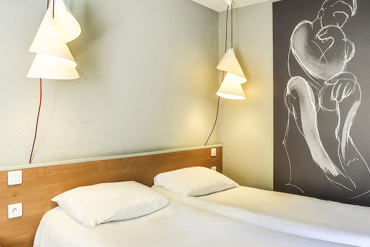 Una habitación de hotel con cabecera de madera, dos camas con ropa blanca y lámparas colgantes.