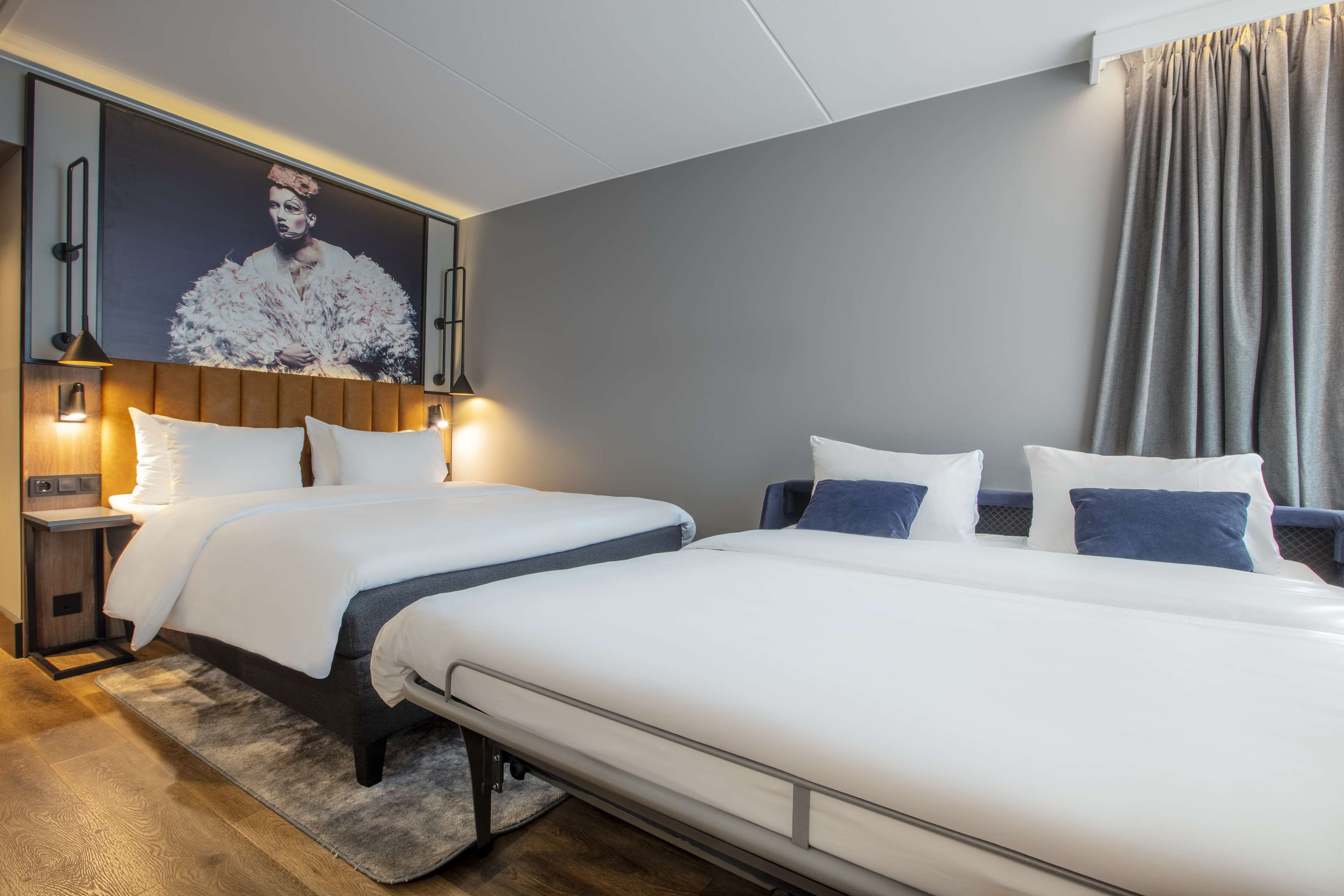 Habitación de hotel con dos camas, una obra de arte grande colgada en la pared, lámparas de noche y cortina gris.
