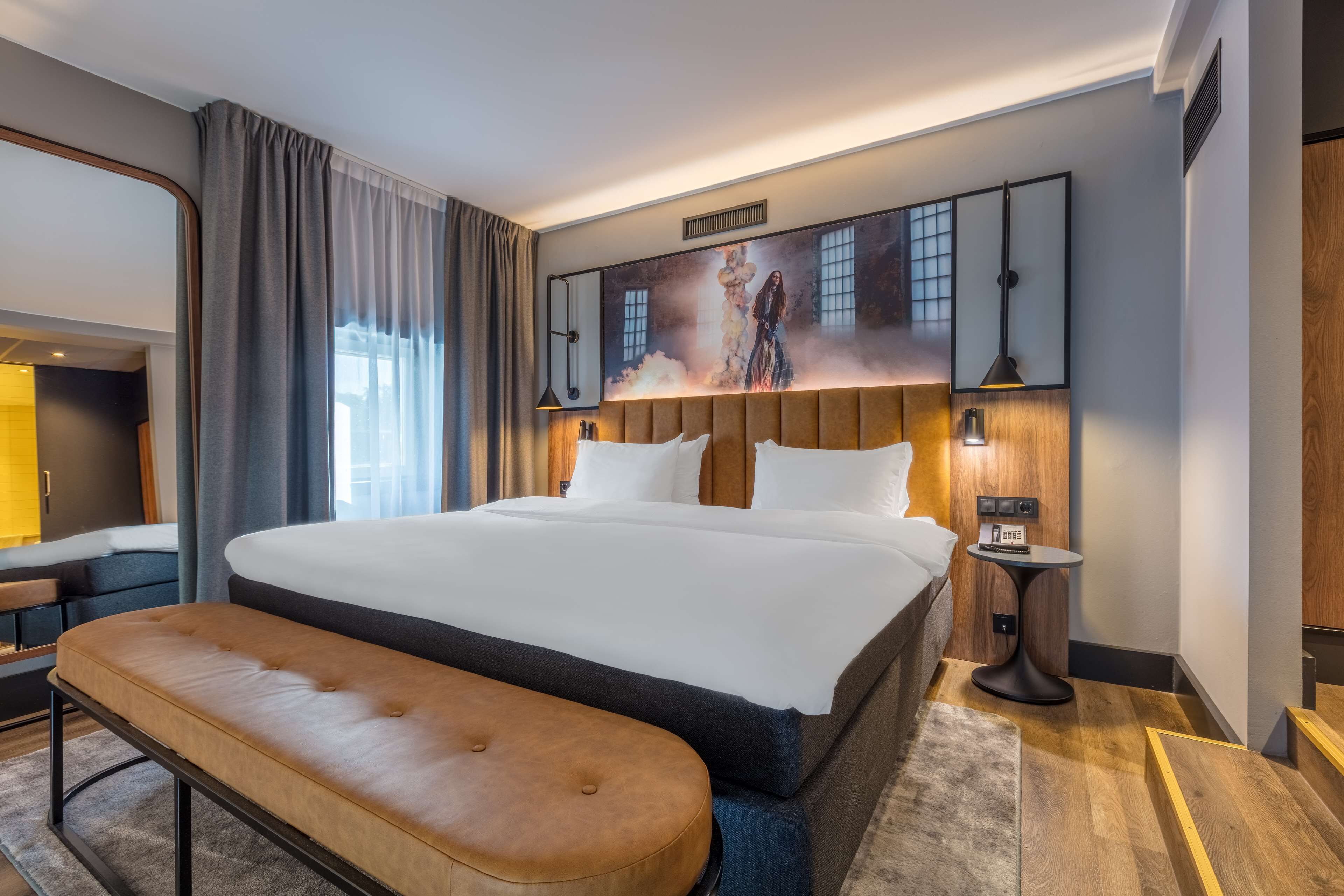 A modern hotel room with a large bed, a bench, a side table, and a large artwork on the wall.