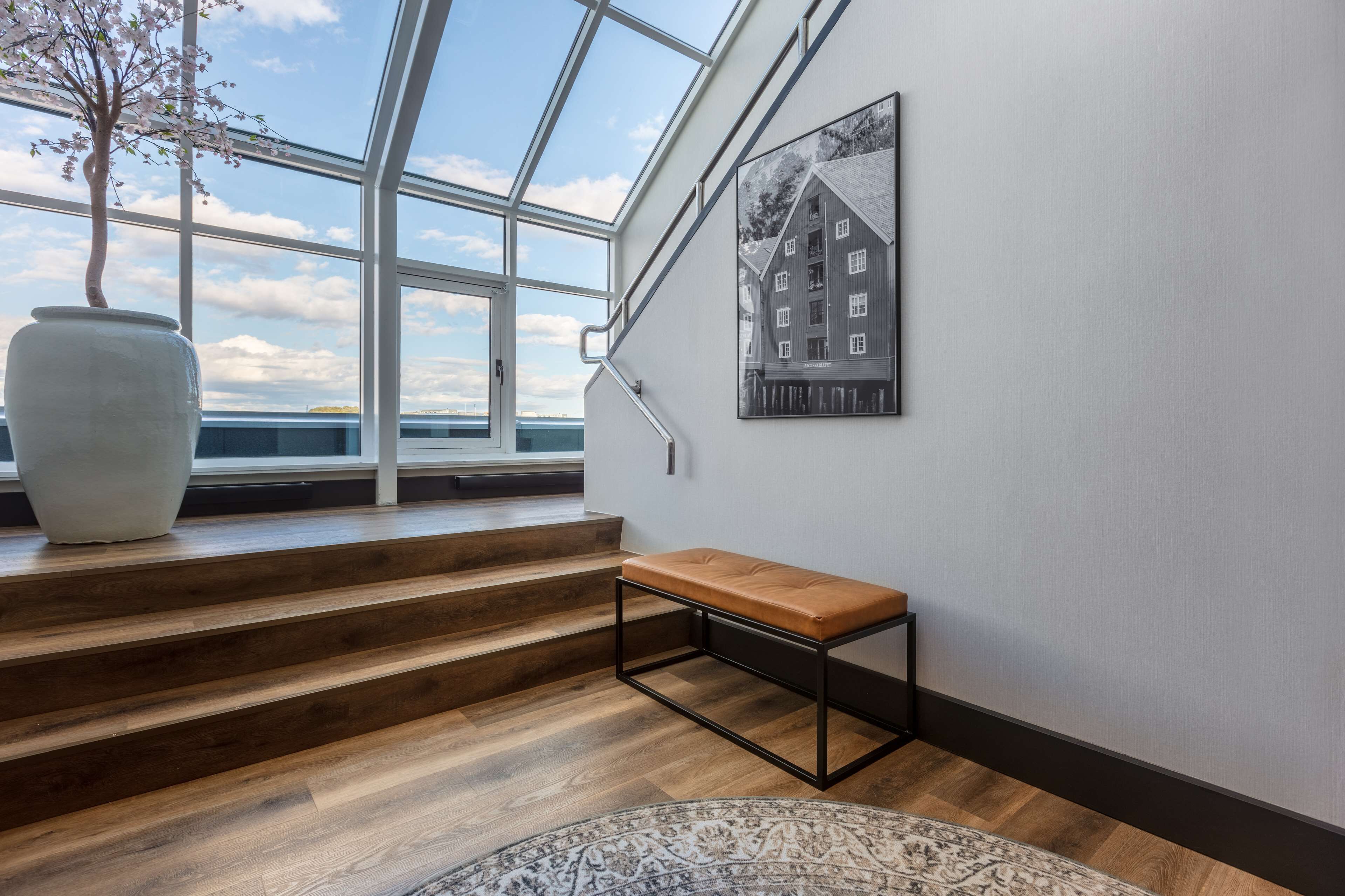 Un escalier avec des marches en bois menant à un palier, un banc, un grand vase et un tableau encadré au mur.