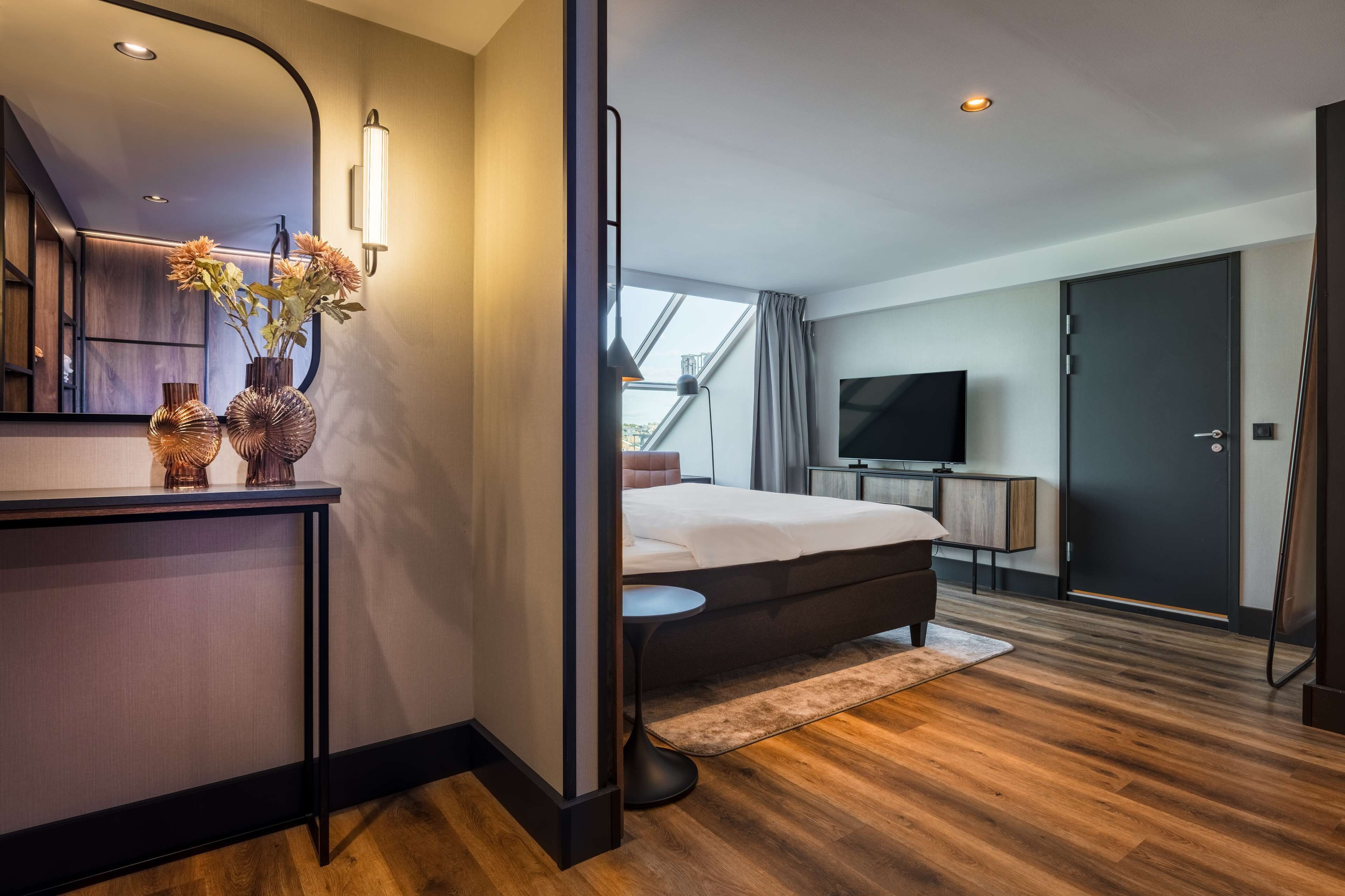 A modern hotel room with a large bed, a TV, a desk with a mirror, and a window with curtains.