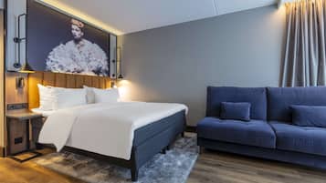 Habitación de hotel moderna con una cama grande, un sofá azul, piso de madera e imagen enmarcadas en la pared.