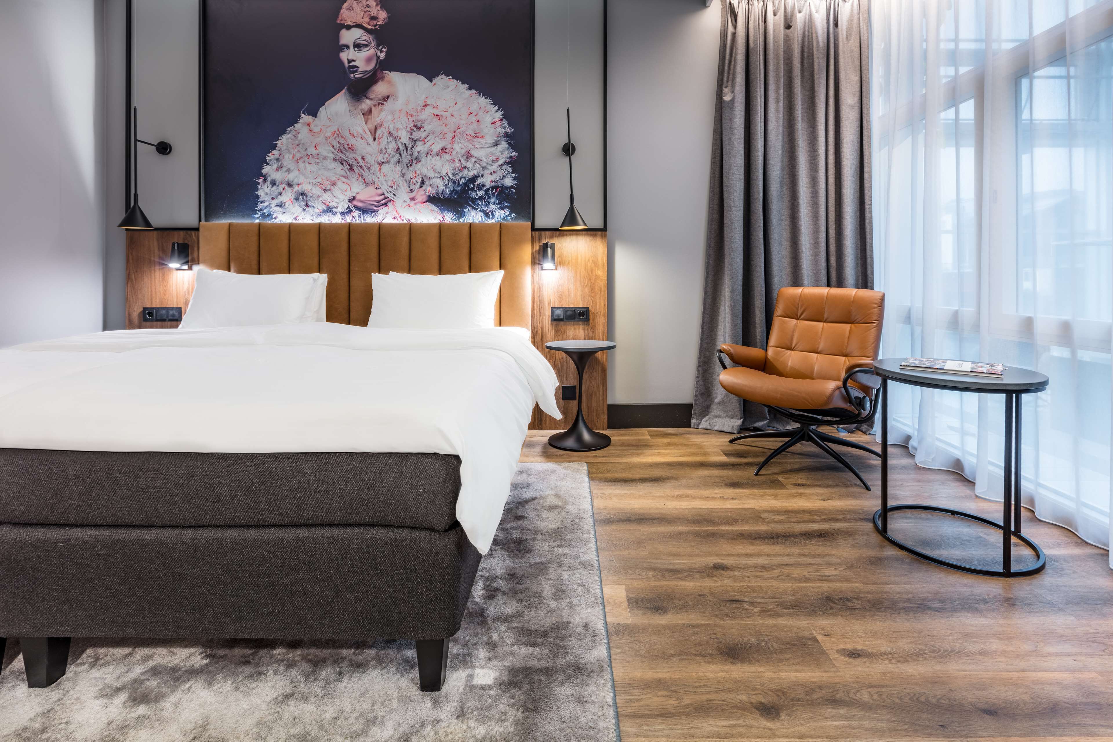 Quarto de hotel moderno com uma cama grande, cabeceira de madeira, área de estar com uma poltrona e uma mesa pequena, e uma janela ampla com cortinas.