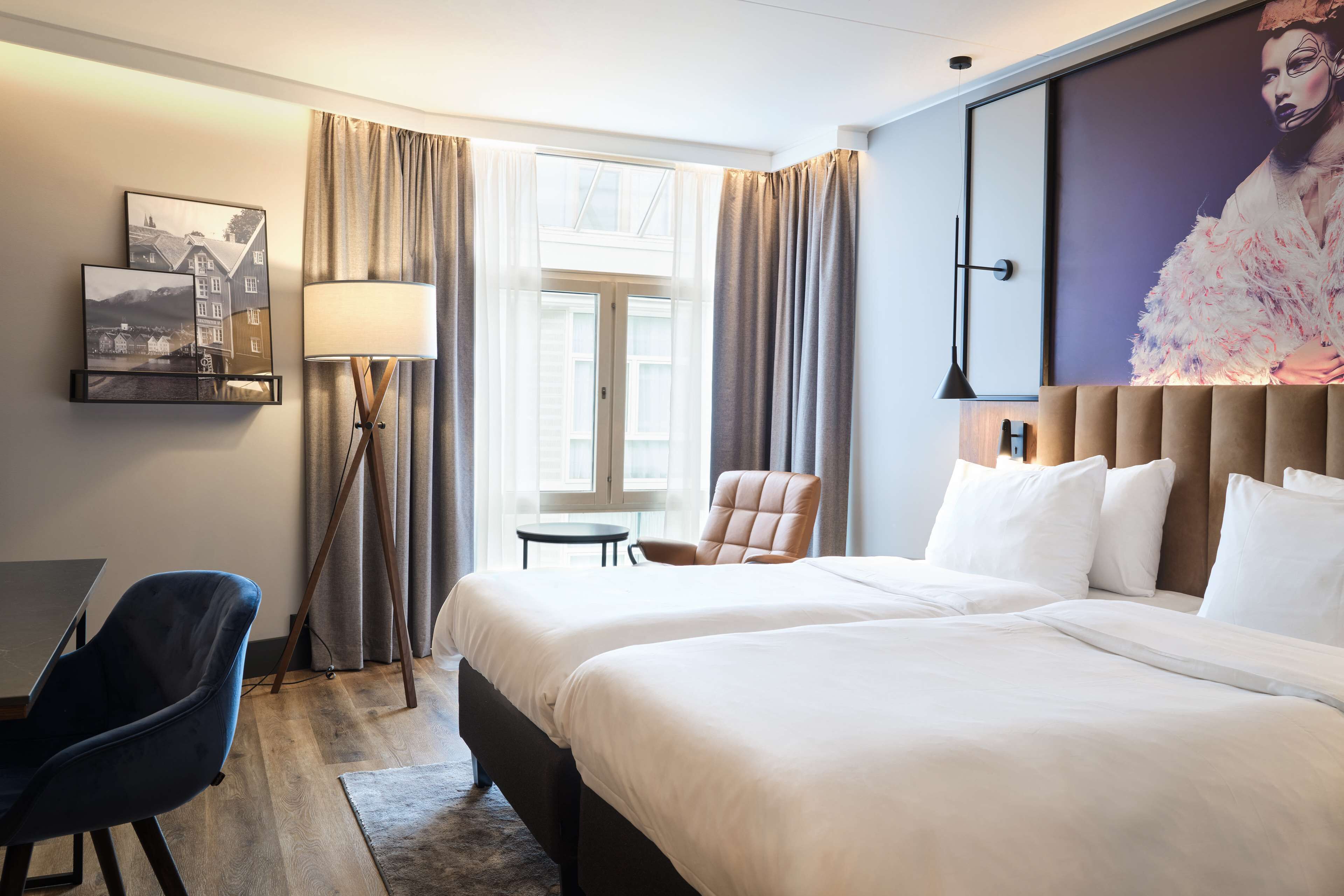 Habitación de hotel con dos camas, un escritorio, una silla, una lámpara y un gran ventanal con cortinas.