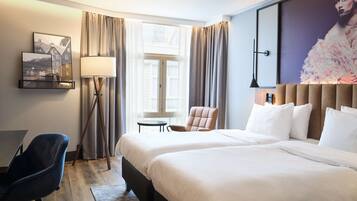 Ein Hotelzimmer mit zwei Betten, einem Schreibtisch, einem Stuhl, einer Lampe und einem großen Fenster mit Vorhängen.