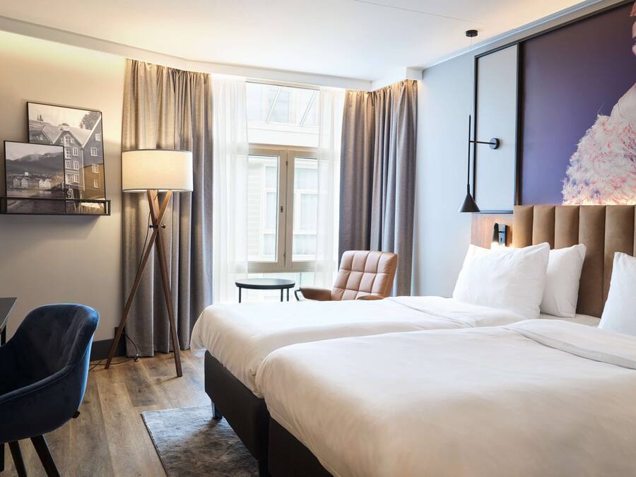 Ein Hotelzimmer mit zwei Betten, einem Schreibtisch, einem Stuhl, einer Lampe und einem großen Fenster mit Vorhängen.
