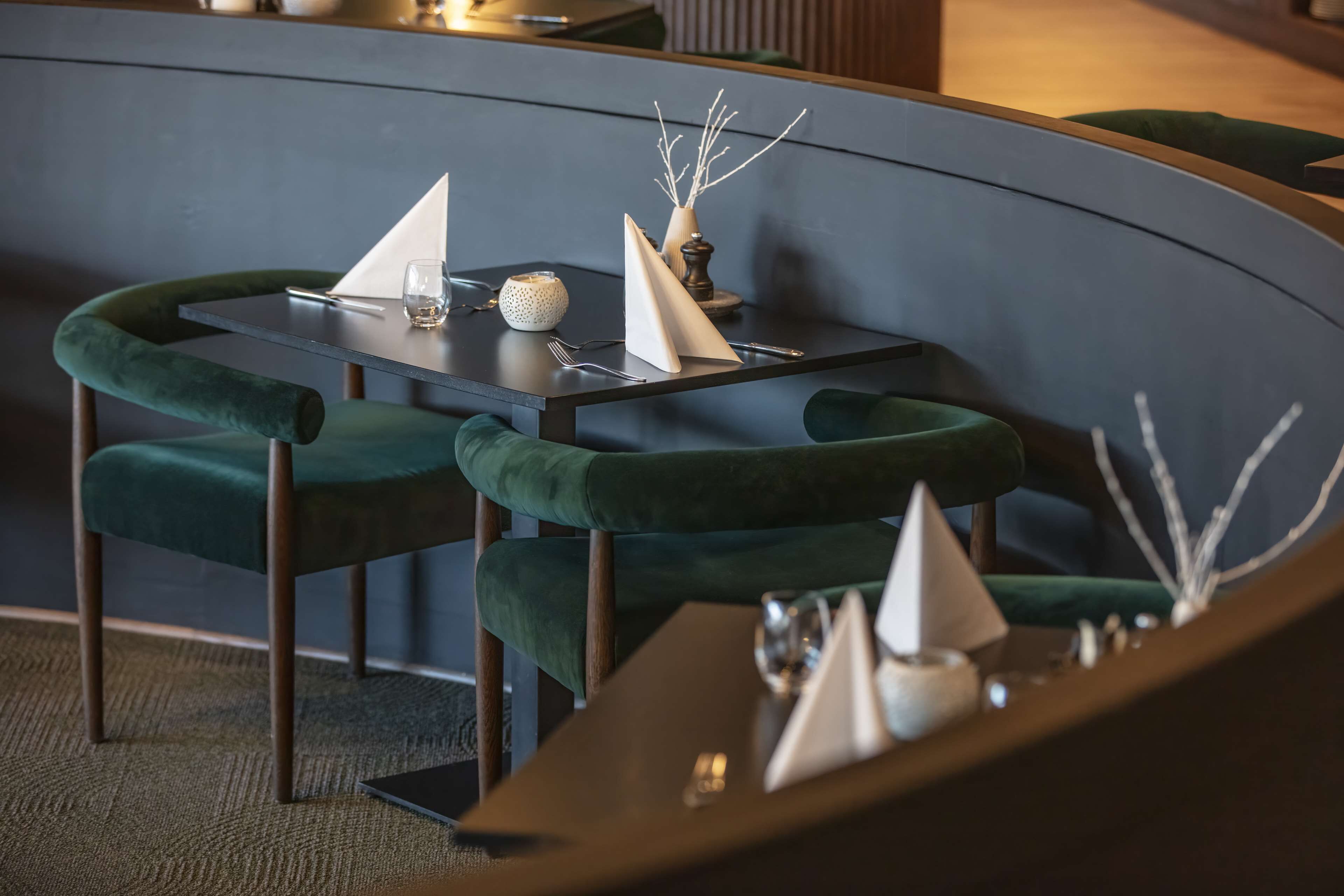 A modern dining area with a curved wall, green upholstered chairs, a dark table, and a small decorative vase with branches.