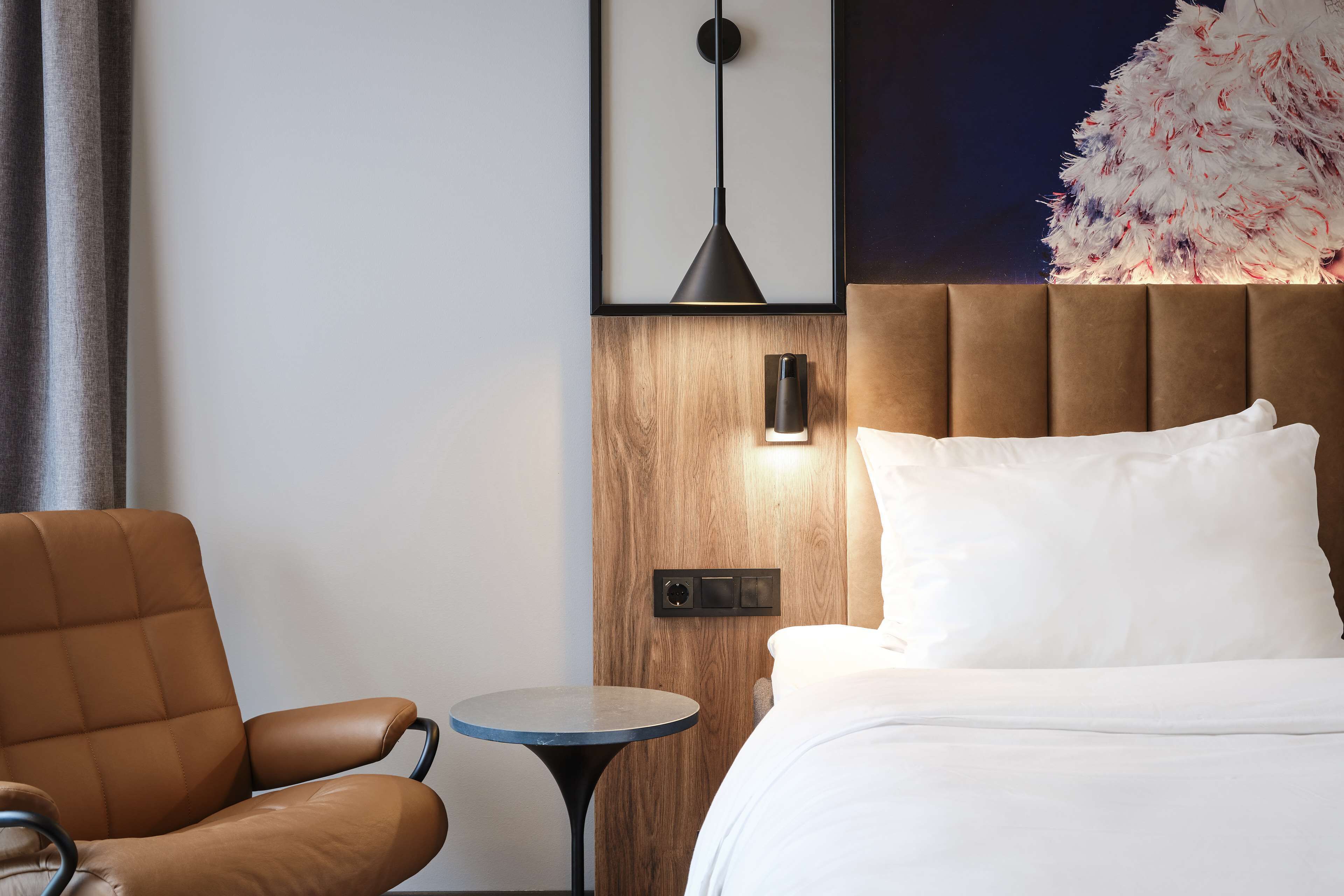 A hotel room with a brown leather chair, a round table, a bed with white bedding, a wooden headboard, and a wall-mounted lamp.