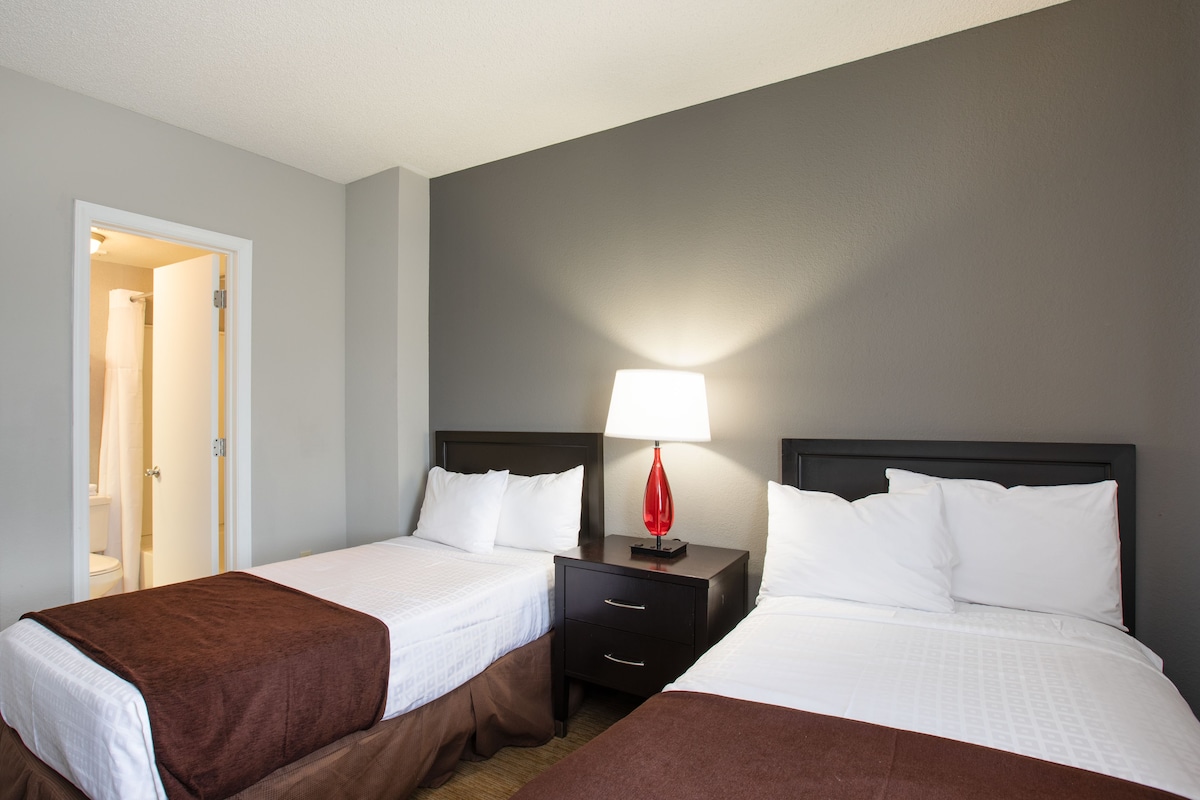 A hotel room with two beds, a nightstand with a lamp, and a door leading to a bathroom.