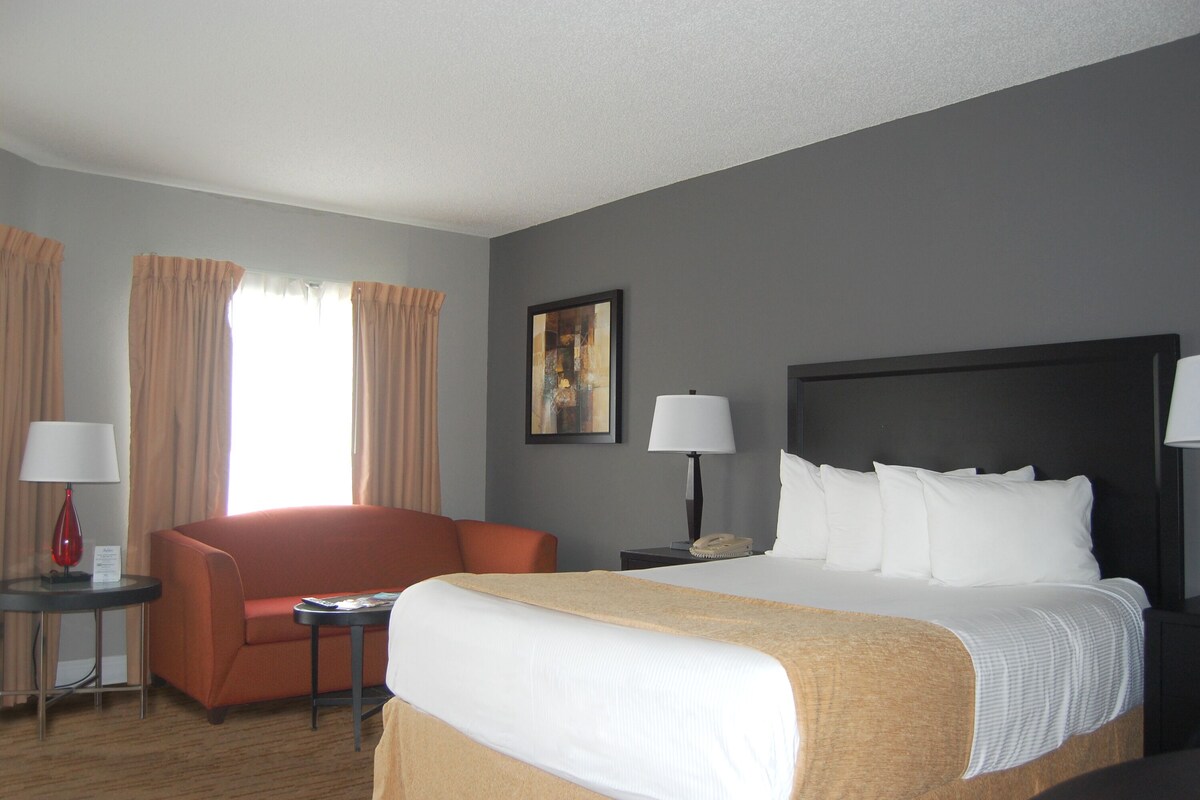 A hotel room with a large bed, a sofa, a desk with a lamp, a telephone, and a painting on the wall.