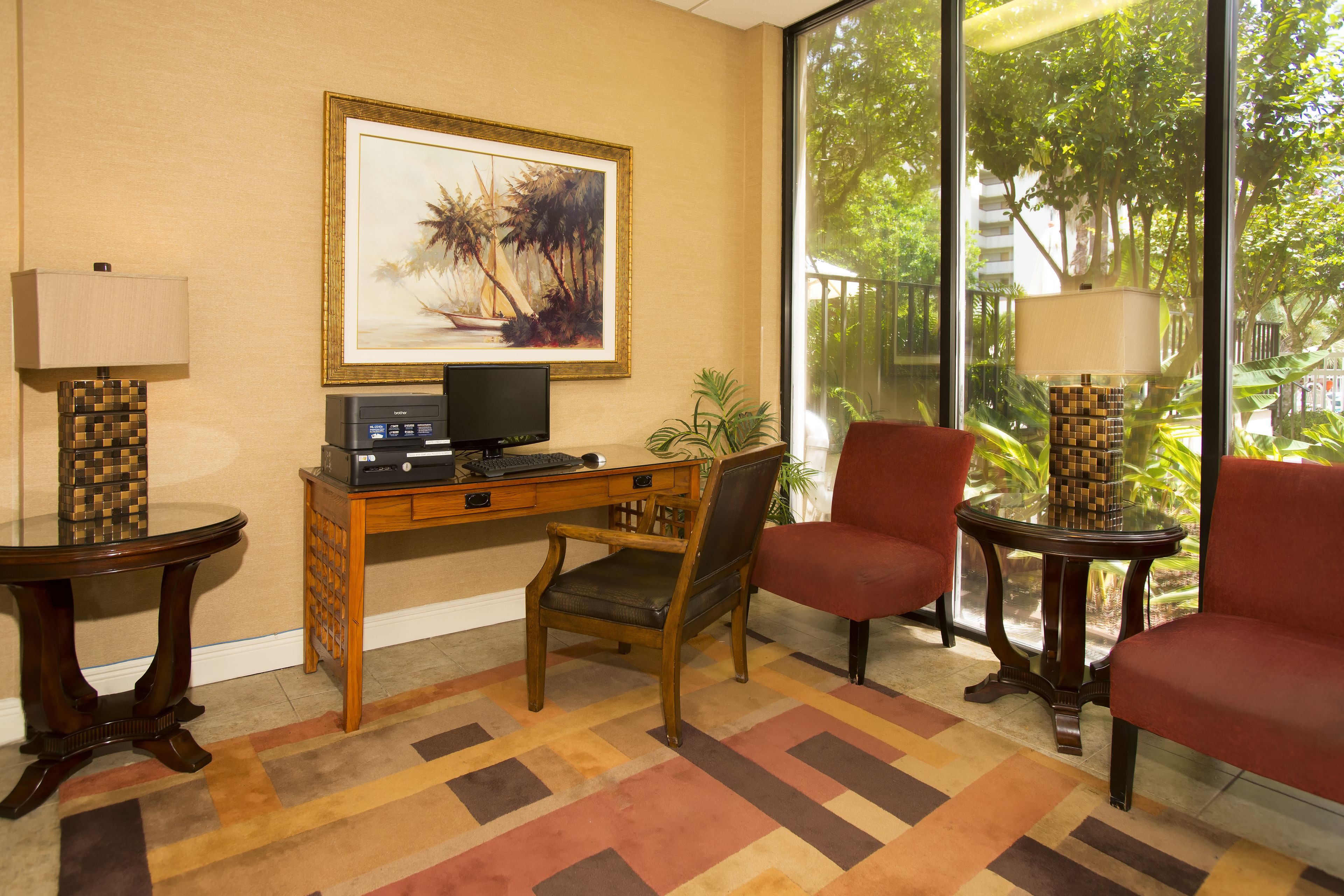 A hotel room with a wooden desk, a computer, a chair, a red chair, a lamp, and a large window with a view of trees.