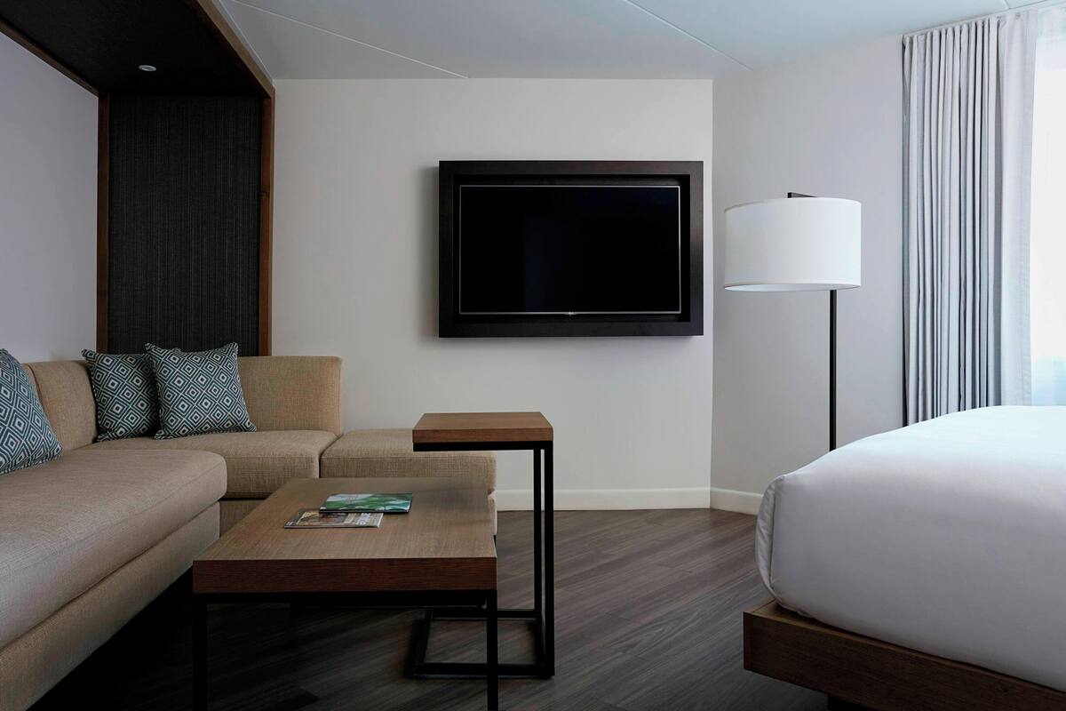 A modern hotel room with a sofa, a small wooden table, a bed, a flat-screen TV mounted on the wall, and a floor lamp.