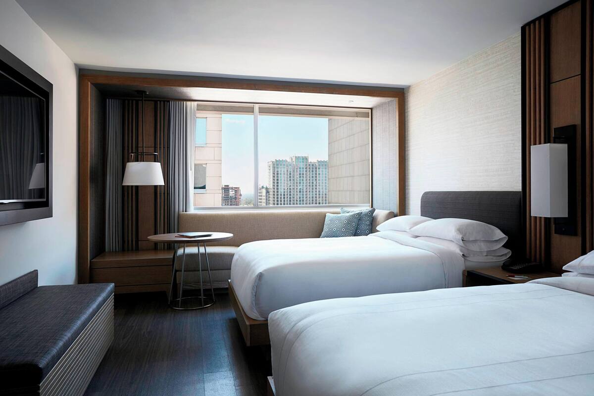 A hotel room with two beds, a small table, a sofa, and a large window offering a city view.
