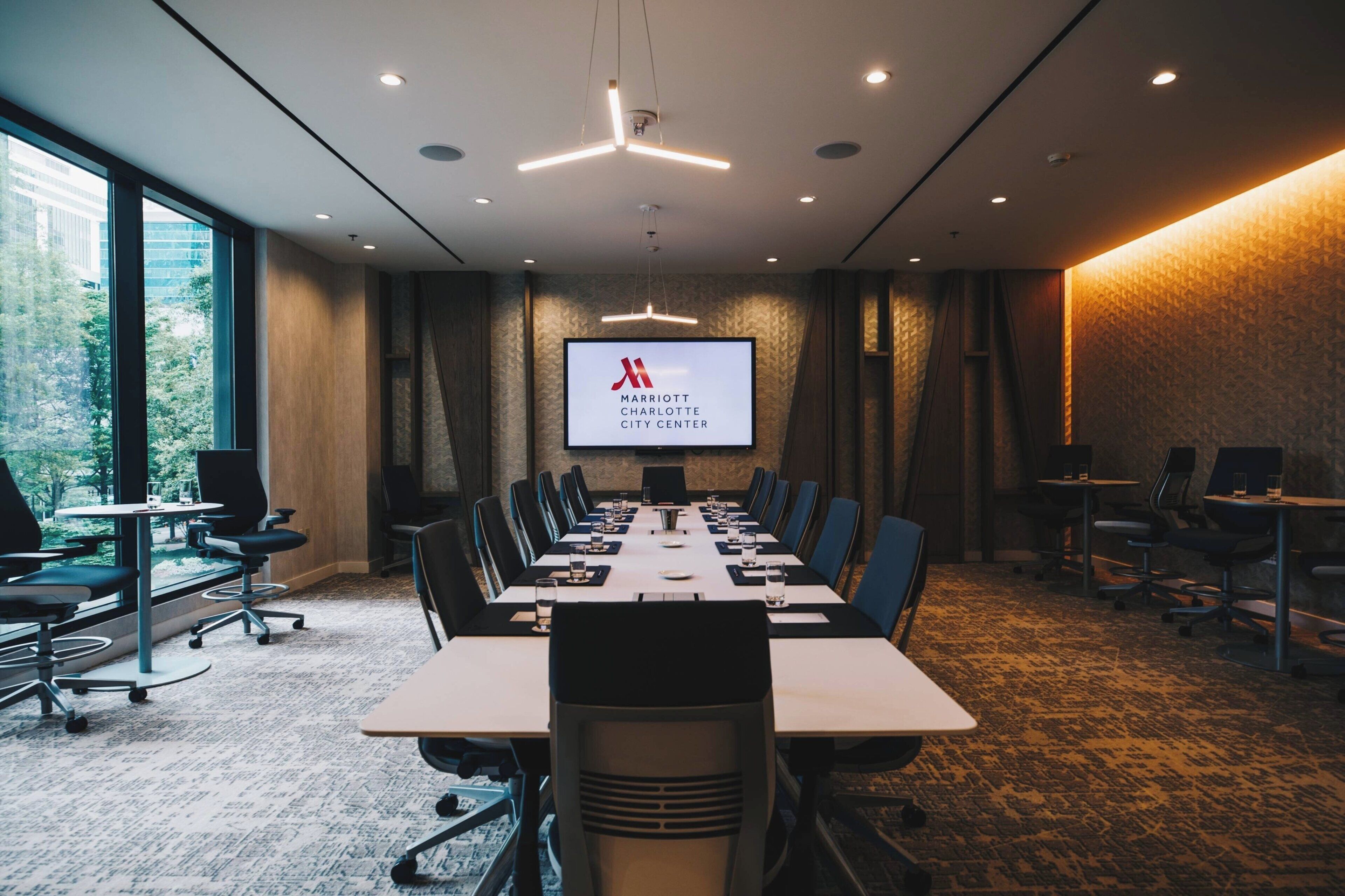 A conference room with a long table, chairs, a projector screen, and a view of trees through large windows.