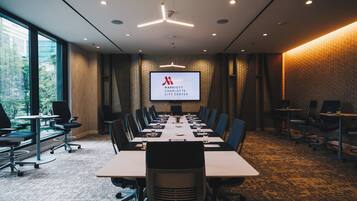 A conference room with a long table, chairs, a projector screen, and a view of trees through large windows.