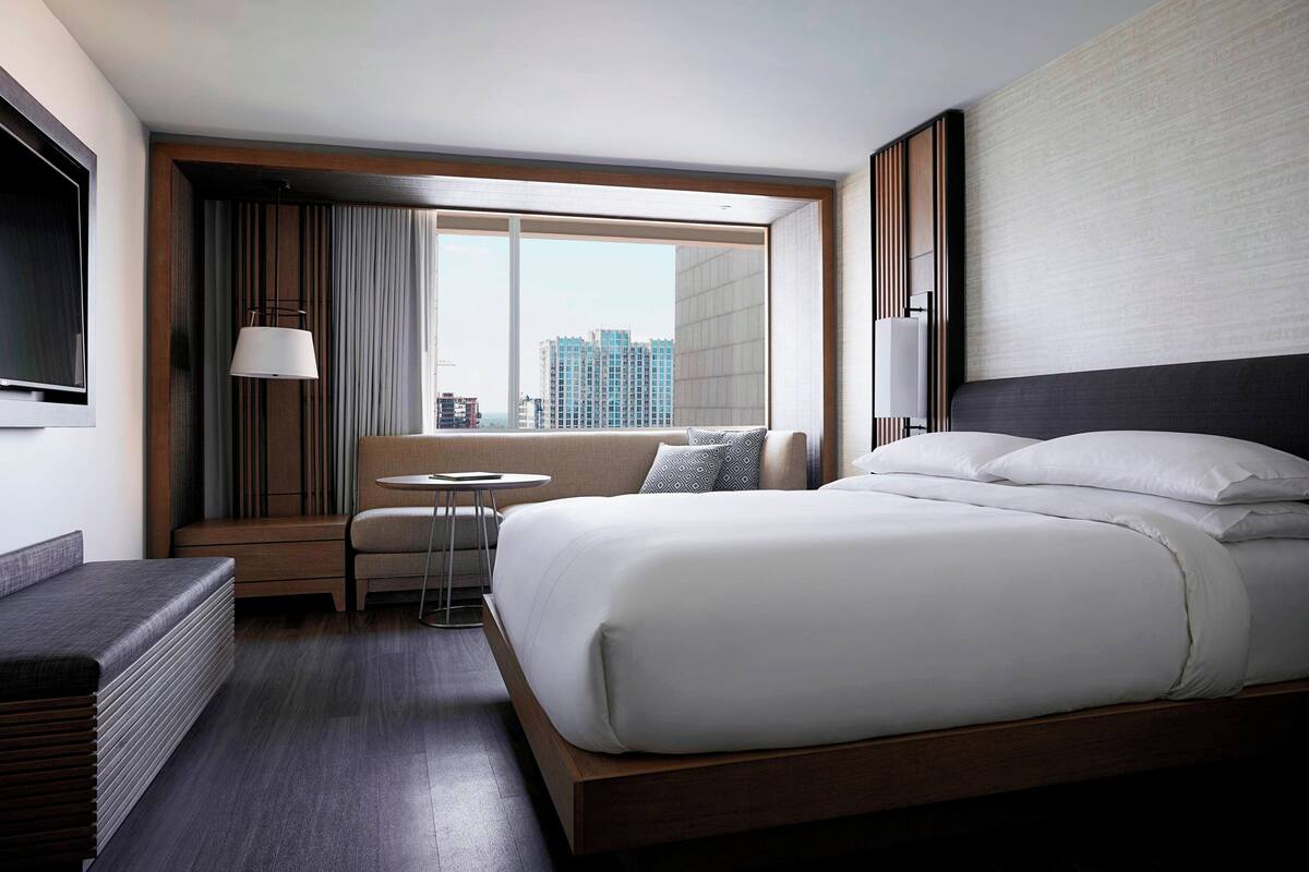 A modern hotel room with a large bed, a small desk, a chair, and a window with a city view.