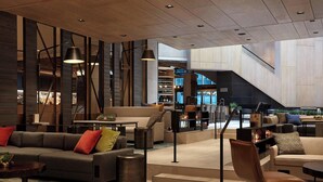 A spacious lobby with a modern design, featuring a large sofa, a coffee table, and a seating area with chairs. There is a staircase leading to an upper level, and a bar area with shelves of bottles. - Charlotte Marriott City Center (Charlotte)