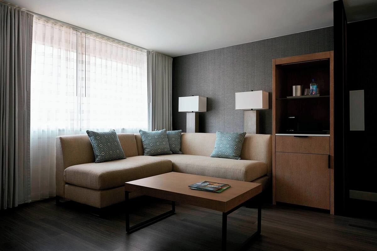A modern hotel room with a beige sofa, a wooden coffee table, and a dark wood floor. There is a large window with sheer curtains, a wall-mounted TV, and a small wooden cabinet.