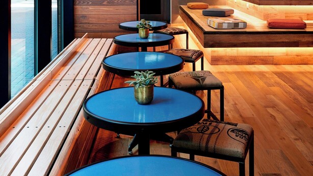 A modern interior with wooden benches and round tables, a kitchen area with a chalkboard menu, and a view of the street outside. - Charlotte Marriott City Center (Charlotte)