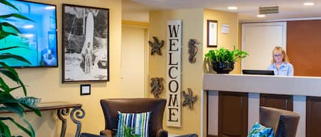 Zona de recepción con un cartel que dice 'WELCOME', una recepcionista y dos sillones con cojines decorativos.