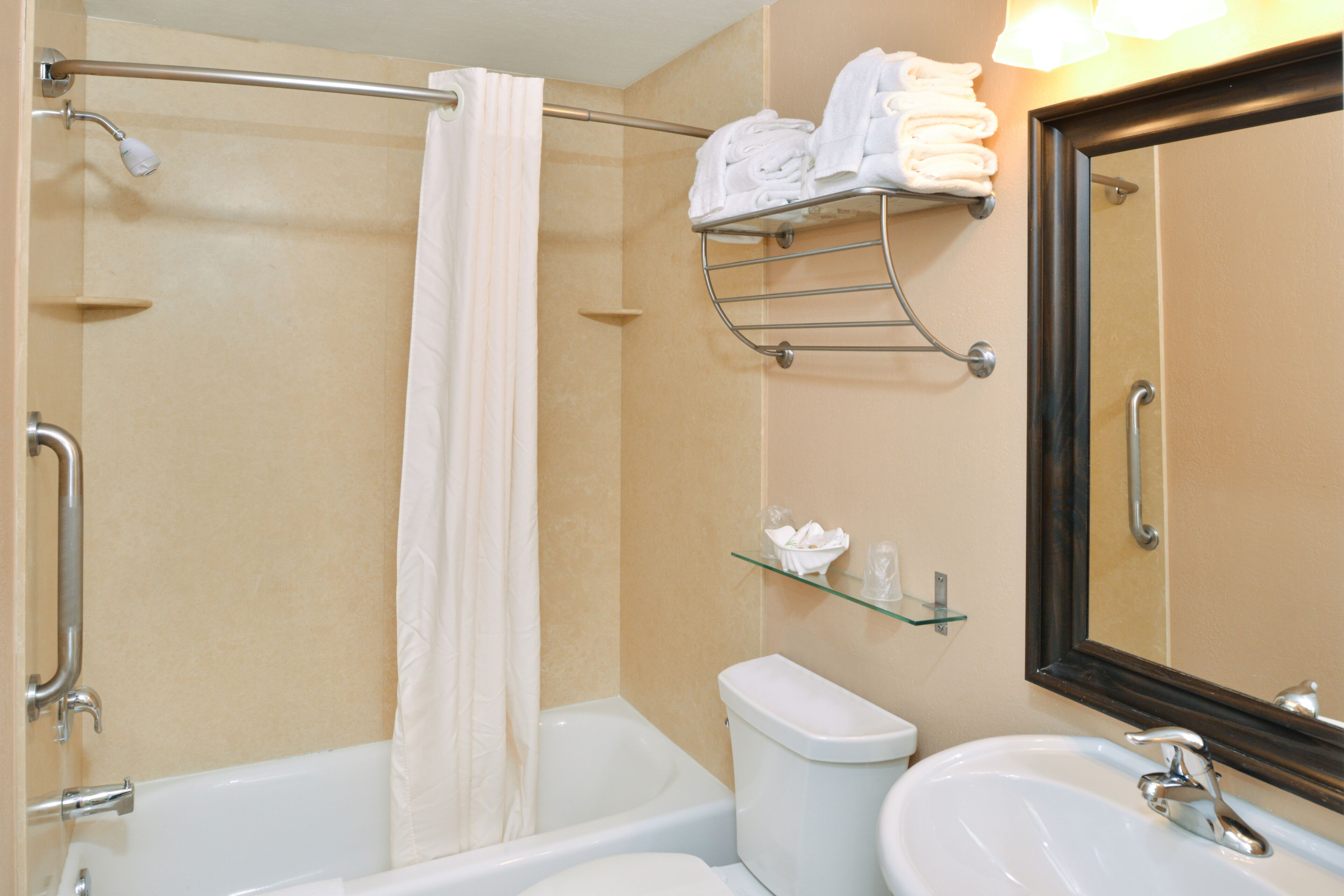 A bathroom with a bathtub, toilet, and sink. There is a shower curtain, a towel rack with folded towels, a glass shelf with toiletries, and a large mirror.