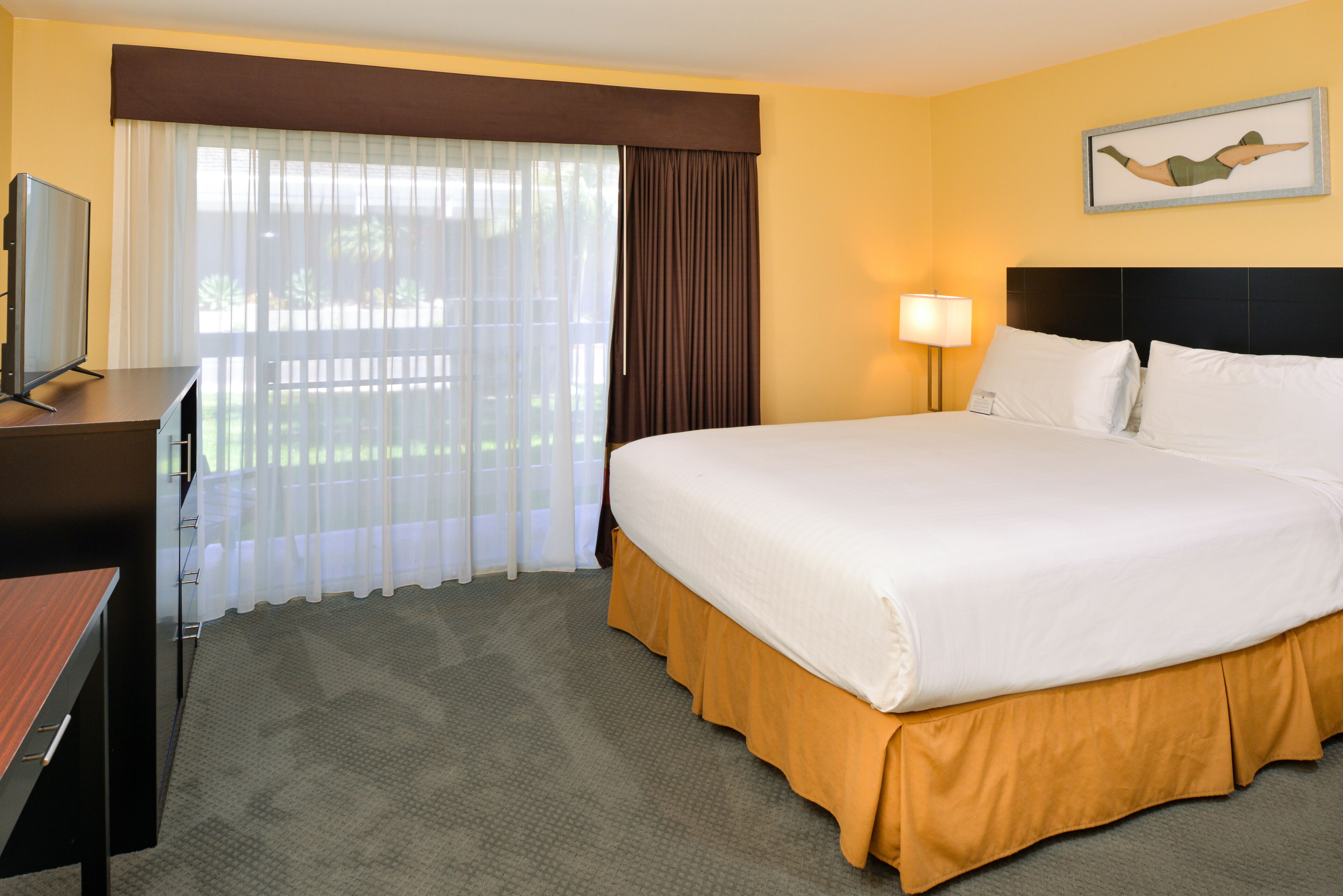 A hotel room with a large bed, a desk with a television, a window with curtains, and a painting on the wall.