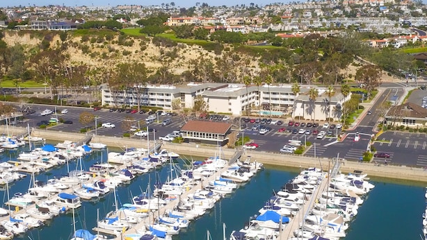 Un port de plaisance où sont amarrés de nombreux bateaux, un parking et, au fond, un immeuble à plusieurs étages.