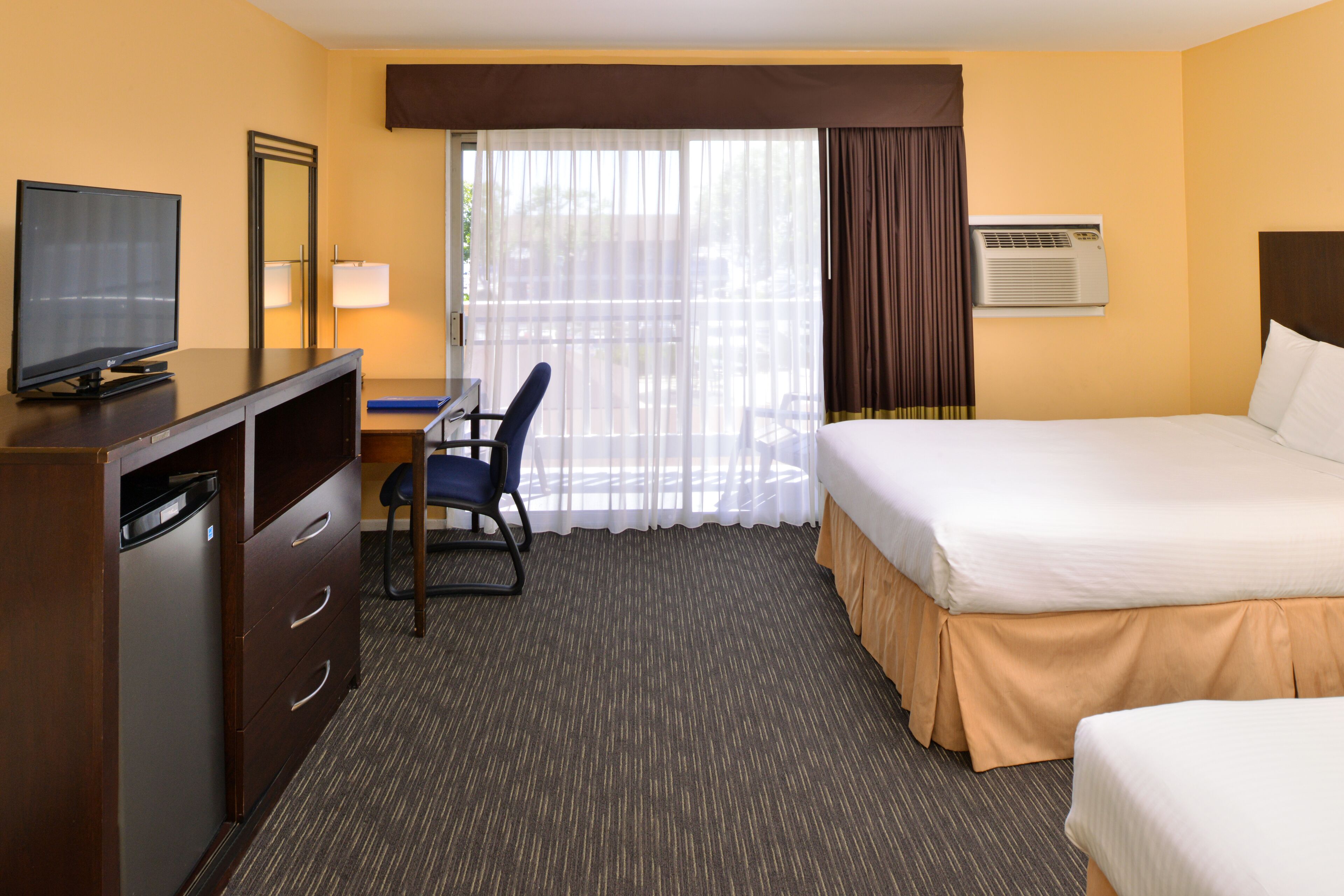 A hotel room with a bed, a desk with a chair, a television, a dresser, and an air conditioning unit.