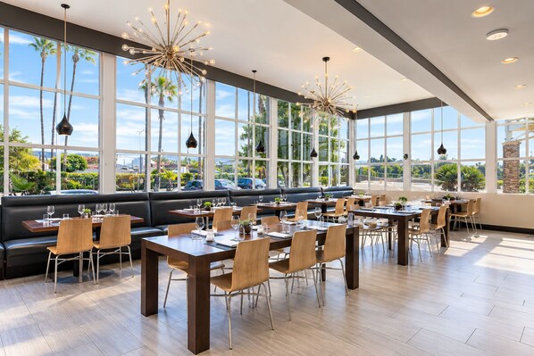 Un restaurante con vistas a palmeras y un cielo despejado. Las mesas están listas con sillas y copas de vino.