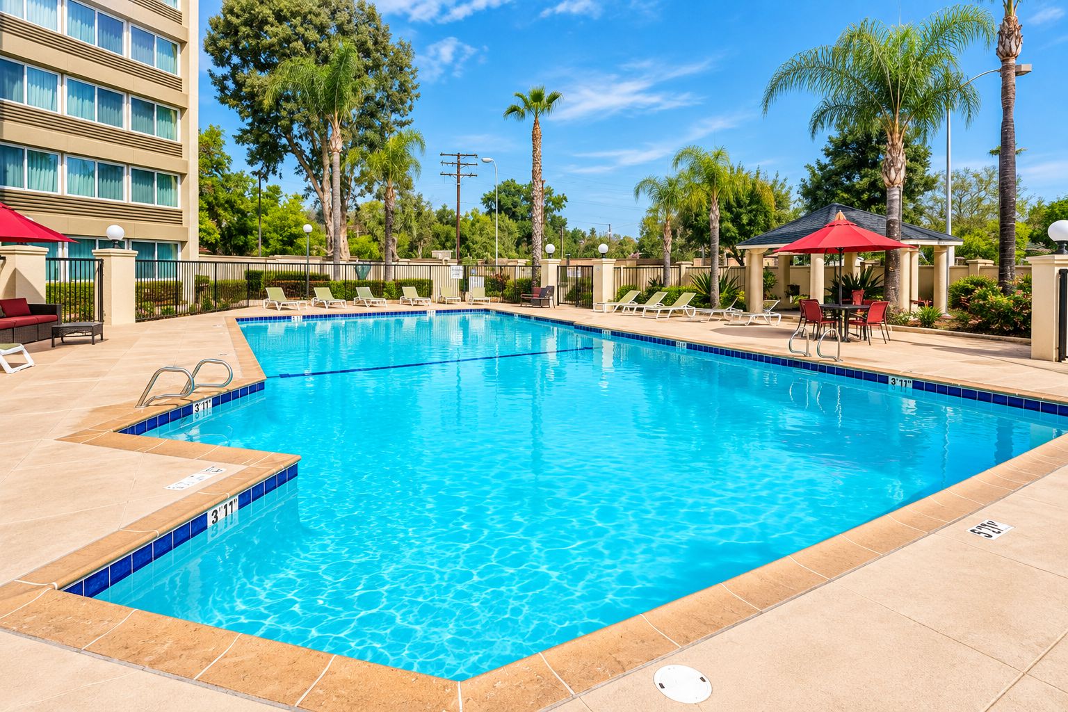 A rectangular swimming pool with clear blue water, surrounded by lounge chairs and umbrellas, with a building and palm trees in the background.