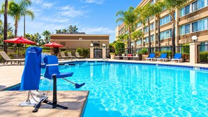 A swimming pool with lounge chairs and umbrellas, surrounded by palm trees and a building with multiple windows. - Pacific Stay Hotel (West Covina)