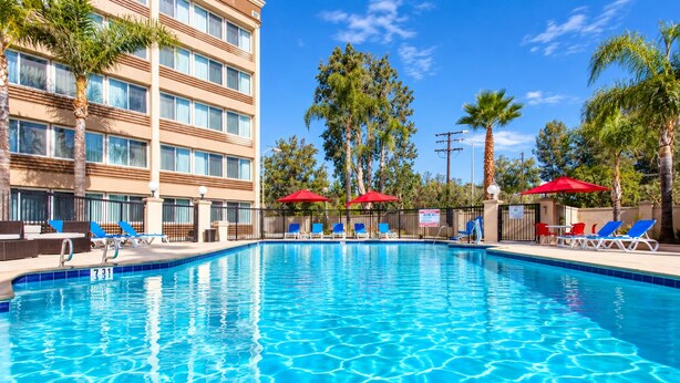 A swimming pool with lounge chairs and umbrellas, surrounded by palm trees and a hotel building. - Pacific Stay Hotel (West Covina)