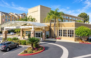 Entrada de hotel con galería techada, palmeras y estacionamiento.