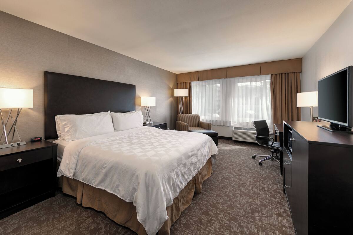 A hotel room with a large bed, two bedside tables with lamps, a desk with a television, a chair, and a window with curtains.