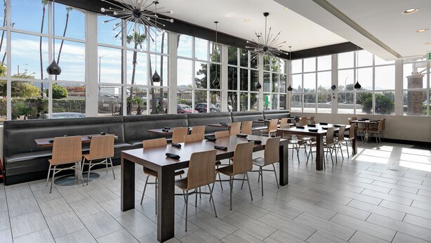A modern dining area with large windows, a black leather couch, and wooden tables and chairs. - Pacific Stay Hotel (West Covina)