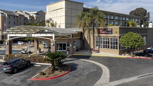 A hotel with a modern facade, a covered entrance, and a parking lot with a red curb. - Pacific Stay Hotel (West Covina)