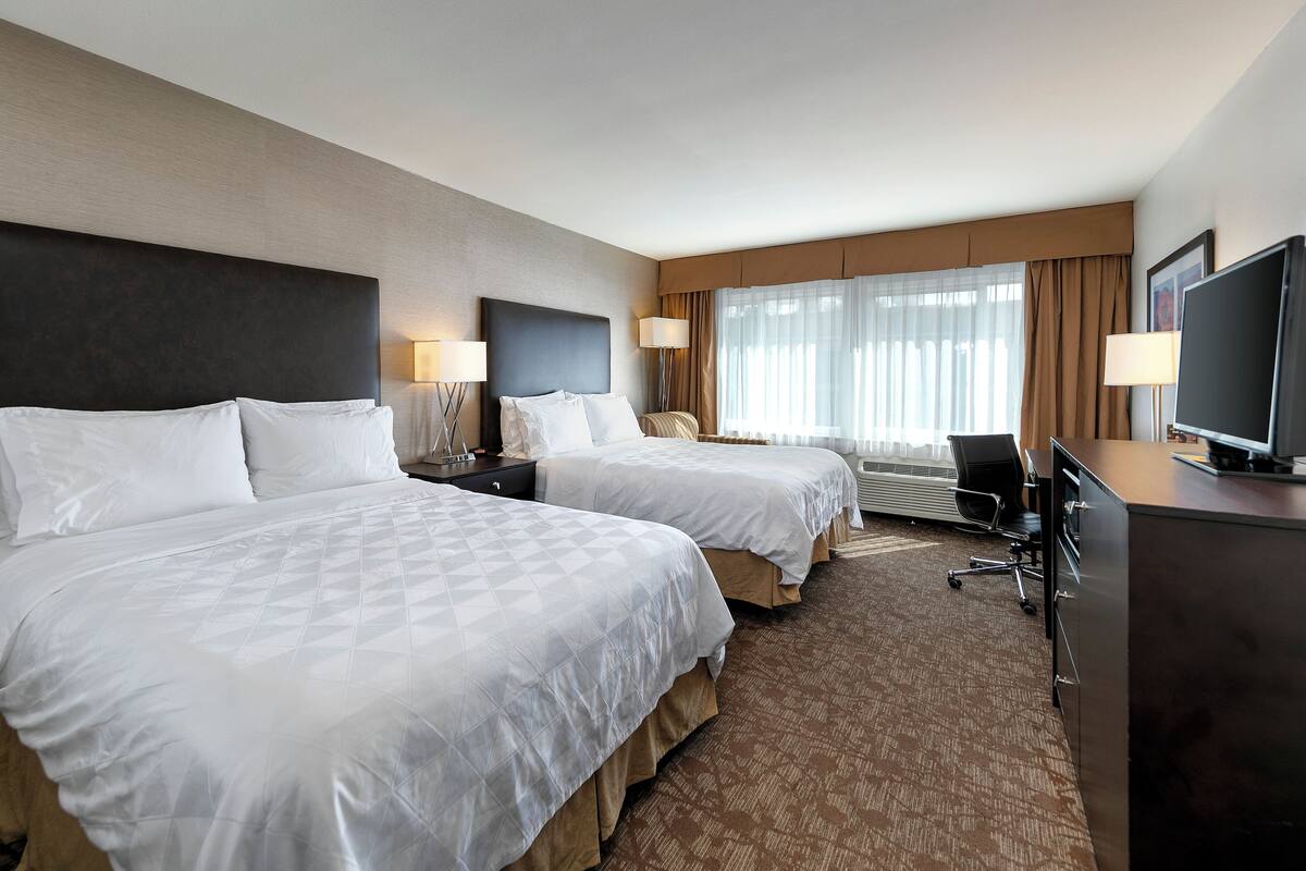 A hotel room with two beds, a desk with a computer, and a large window with curtains.