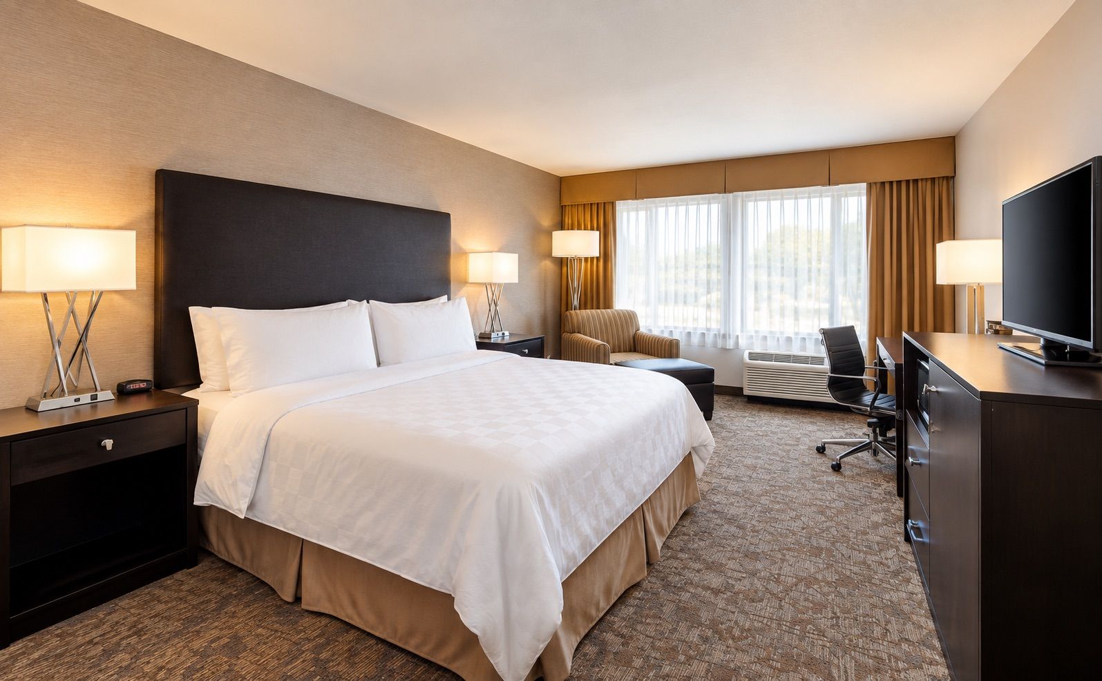 A hotel room with a large bed, two bedside tables with lamps, a desk with a chair, a television, and a window with curtains.