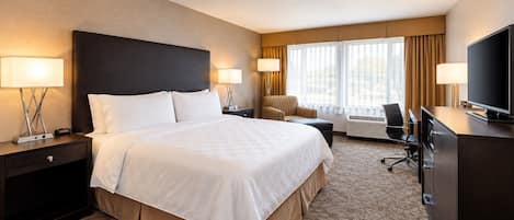 A hotel room with a large bed, two bedside tables with lamps, a desk with a chair, a television, and a window with curtains.
