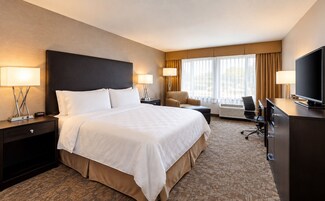 A hotel room with a large bed, two bedside tables with lamps, a desk with a chair, a television, and a window with curtains.