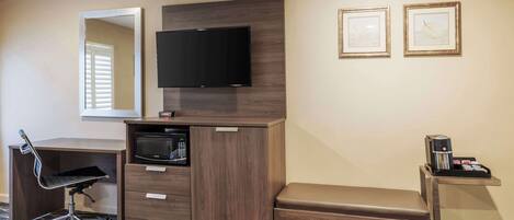 A hotel room with a desk, a microwave, a bench, a TV, and a coffee machine.