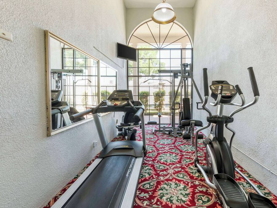 A narrow hallway with a treadmill, elliptical, and stationary bike, carpeted floor, and a large mirror on the left.