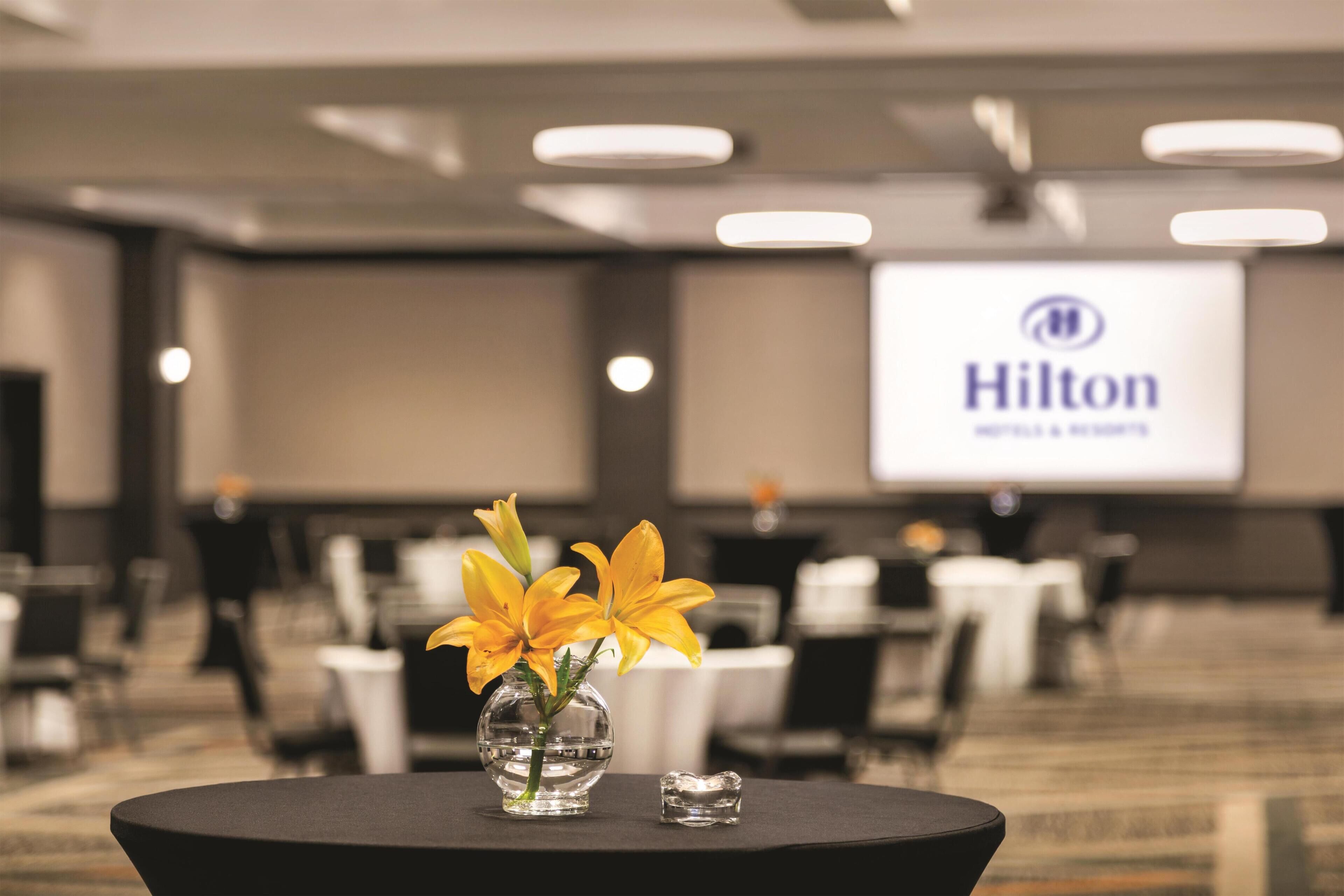 A conference room with a Hilton logo on the screen, a table with a vase of yellow flowers, and a small glass object.