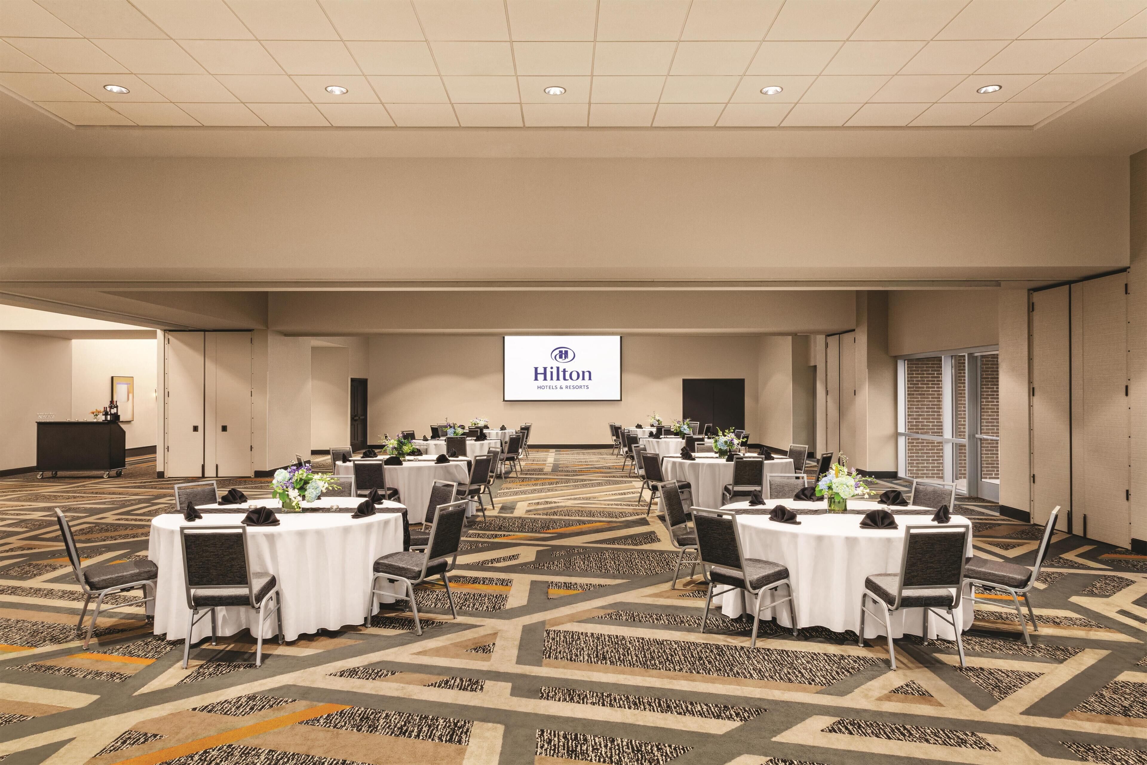 A large, well-lit conference room with multiple round tables, each with chairs and centerpieces. A large Hilton logo is visible on the wall.