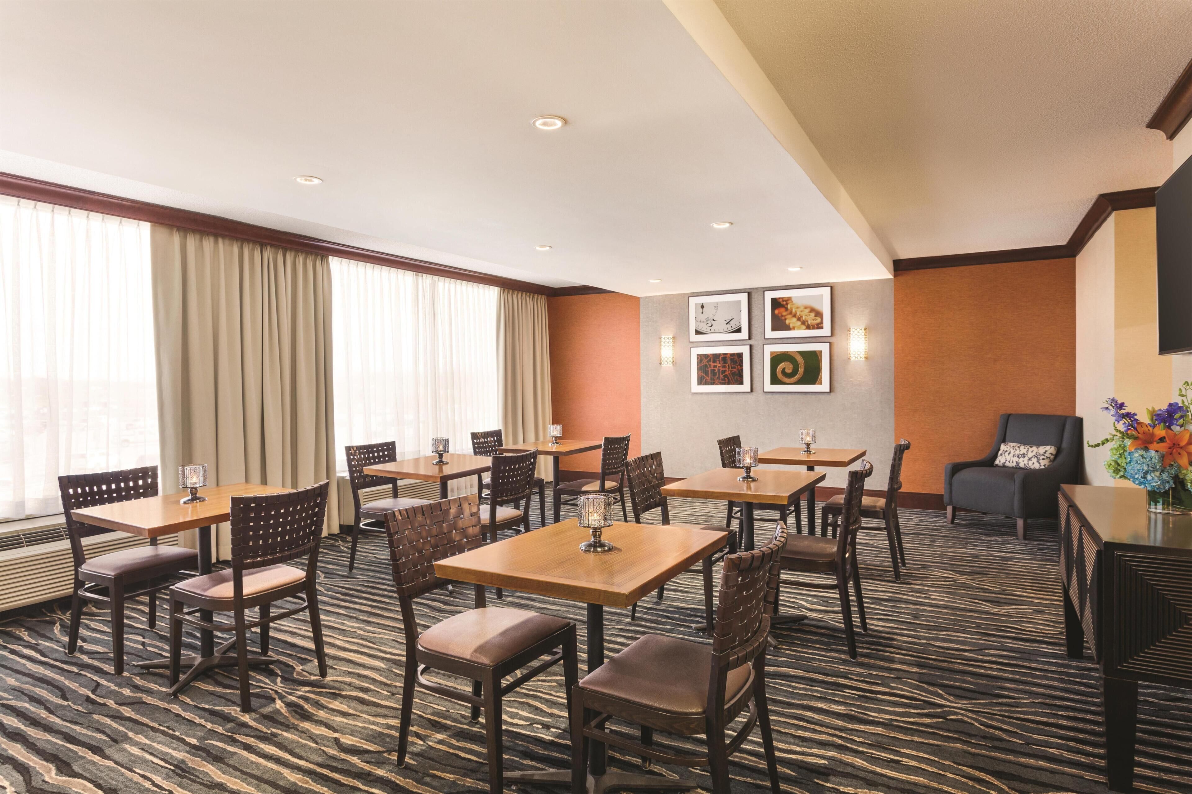 A hotel dining area with multiple tables and chairs, a patterned carpet, and framed artwork on the wall.